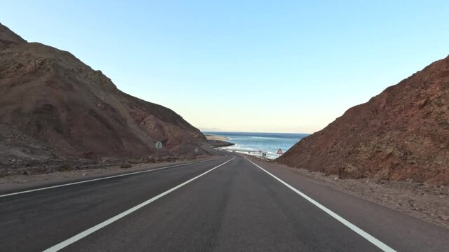 Car driving on desert highway between rocky mountains near sea at sunset
