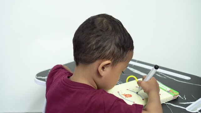 Asian little boy is learning to color with black marker on a worksheet book for pre-school learning                                             