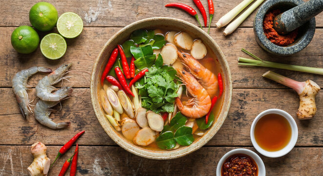 An overhead "flat lay" photograph of a large bowl of clear-broth Tom Yum Goong surrounded by its raw ingredients on a worn wooden table. The bowl is packed with bright red chilies, green kaffir lime l