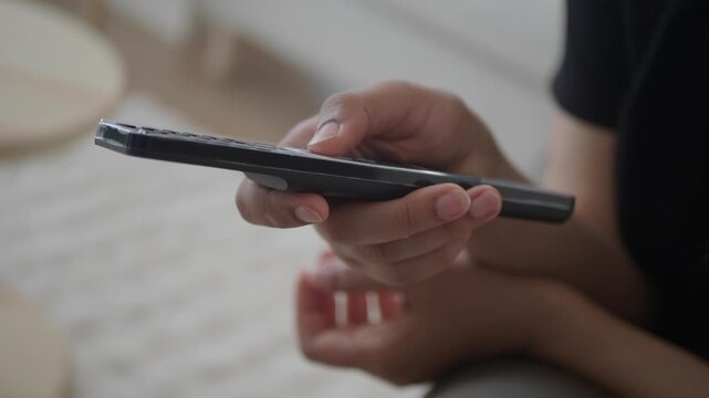 Close up of person's hand pressing a button on a remote control in modern living room, switching channels, changing volume, or navigating smart TV interface