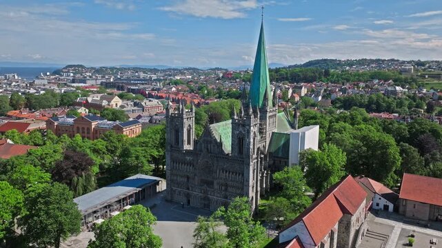 Aerial view of Nidaros Cathedral and Trondheim cityscape, Norway. Drone frames the gothic cathedral with its tall spire rising above trees and red rooftops in sunny weather.