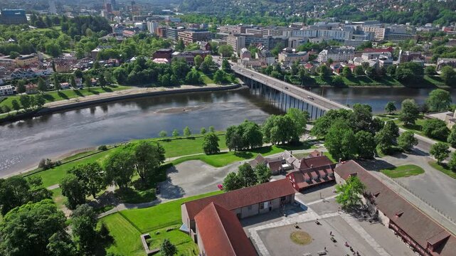 Aerial view of Trondheim and the Nidelva River, Norway. Drone pans above a wide bridge, green riverbank park, and city neighborhoods in bright daylight with clear visibility.