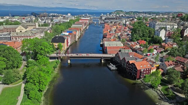 Aerial view of Gamle Bybro over the Nidelva River in Trondheim, Norway. Drone frames the historic bridge and colourful Bakklandet waterfront under bright daylight and clear skies.