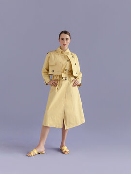 Young beautiful woman with brown hair wearing trendy clothes yellow trench coat and sndals looking confident fresh and lovely Fashion model full length body studio portrait on purple background 