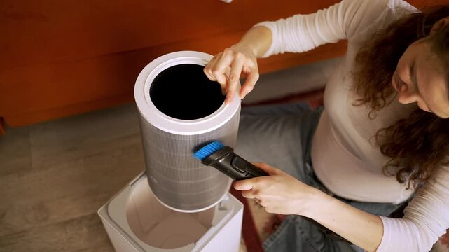 Close-up of a young woman's hands meticulously cleaning a dirty hepa filter from a modern air purifier using a small brush, ensuring clean air and proper appliance maintenance at home