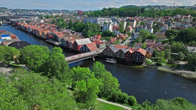 Aerial view of Bakklandet and the Nidelva River in Trondheim, Norway. Drone pans above colorful waterfront buildings and bridges with lush trees in sunny summer light.