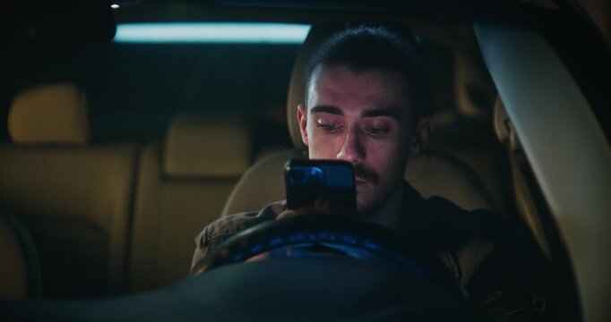 Guy With Tied Back Long Hair Sits in Car, Attentively Reading Messages on Smartphone. Man Pauses Thoughtfully Before Typing Reply, Engaged in Important, Meaningful Conversation. Social Communication.
