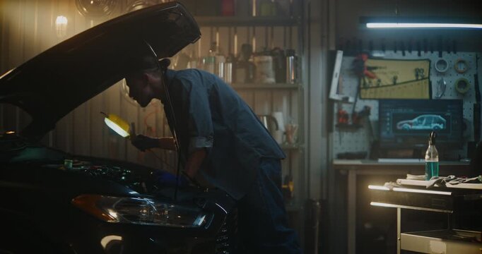 Man Stands in Well Equipped Garage Near Car With Hood Open, Conducting Technical Inspection Under Bright Service Lamp. Guy Examines Engine Components, Ensuring Proper Maintenance, Vehicle Performance.
