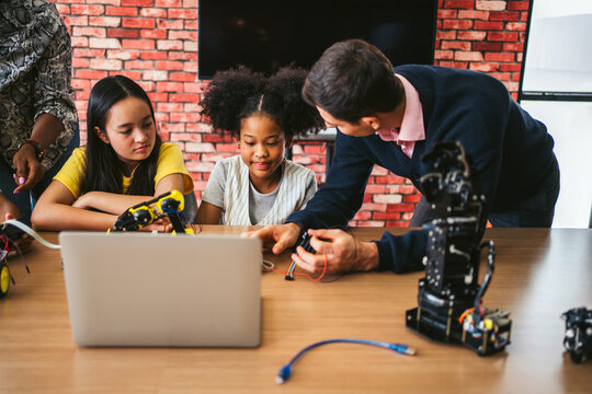 Teacher guides two students building a robot with a laptop in a classroom, surrounded by electronics and parts during a STEM workshop.