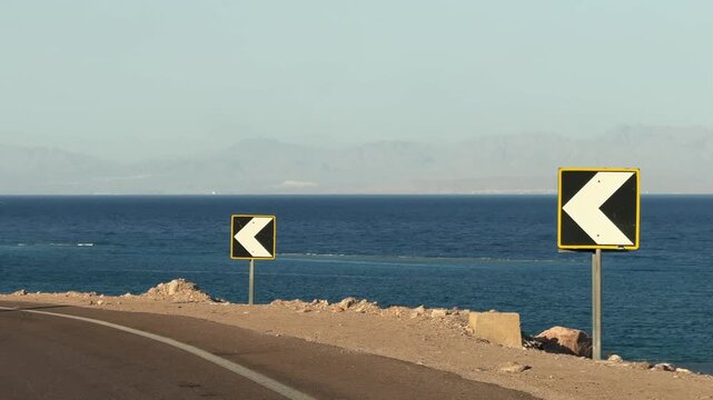 Elevated serpentine desert coastal road turn along the sea with mountains and warning signs
