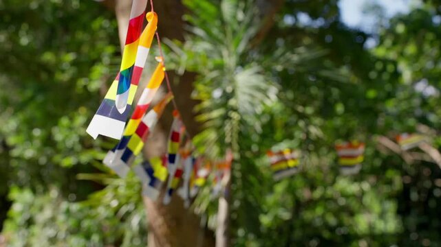 Vibrant multi-colored Buddhist flags hanging on a string, fluttering slightly against a blurred background of green tropical trees.