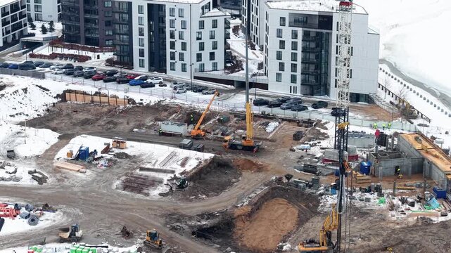 Drone video captures construction work on snowy urban site. Cranes and equipment actively reshape the landscape amid modern high-rise buildings. Cloudy sky adds depth.