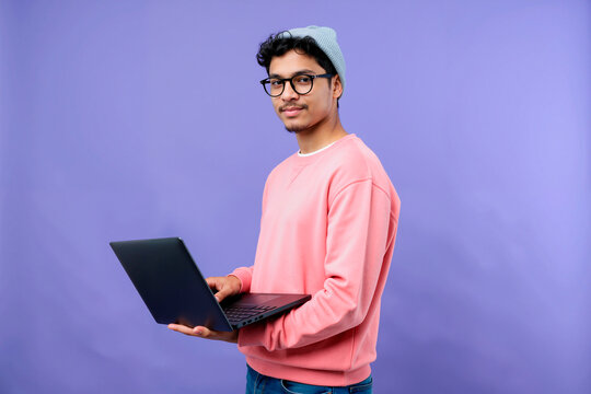 Portrait of young adult Hispanic man wearing glasses looking at camera holding laptop standing against purple background, showing confident expression and casual posture