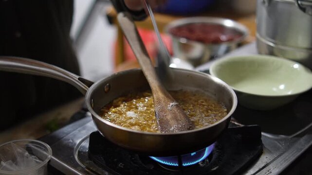Anonymous woman cooking instant noodles or "Mie Nyemek" in a pan, stirring noodles with savory thick sauce and spices in a home kitchen