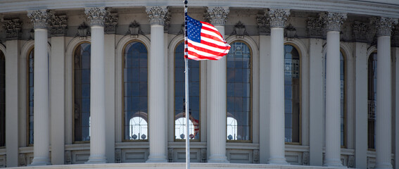 Naklejka premium Capitol with american flag near dome landmark. American flag near capitol with classical columns. Us flag on capitol as political symbol. Capitol with american flag for national background.