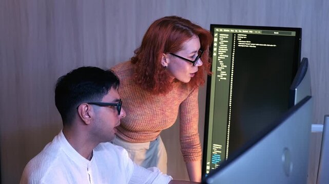 Two colleagues collaborate at a computer, reviewing code on a large monitor in a modern workspace.