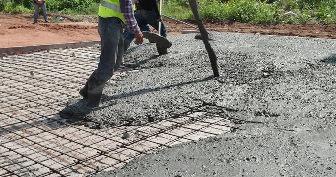 close up of a worker pouring concrete onto a rebar mesh, while other laborers in the background use hoes to spread and level the mixture.