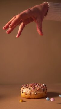 Slow motion of hand releasing golden yeast donut with vanilla glaze and colorful candy prinkles. Donut floats downward against warm beige studio background. Strong visual hook for content and reels.