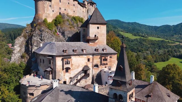 Drone moving near Orava Castle in Slovakia revealing church tower and lower medieval complex with visitors on terrace above green valley.