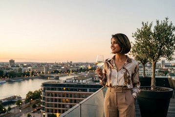 Fototapeta premium A woman in a stylish outfit holds a glass of white wine as she looks out over a city skyline at sunset.