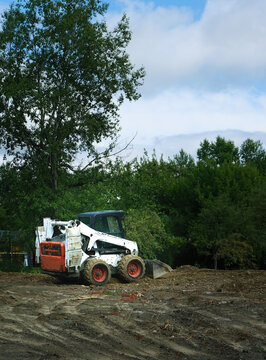 Mini tractor with a bucket at work on a construction site in the public park. Backhoe Loader. Farm and construction equipment, landscaping, Small white and red mechanical digger truck 