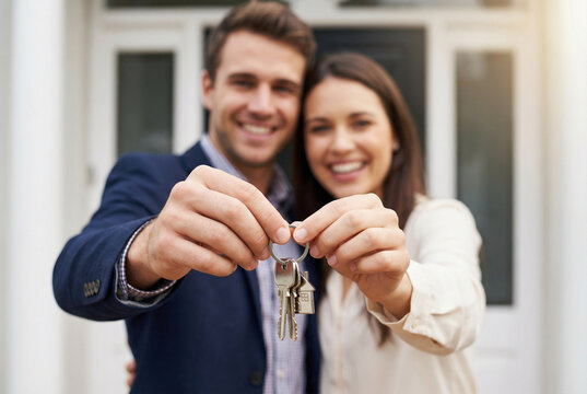 Happy Young Couple Holding Keys to Their New Home, First Time Homeowners Celebrating Real Estate Purchase, Smiling Man and Woman Standing in Front of House Door