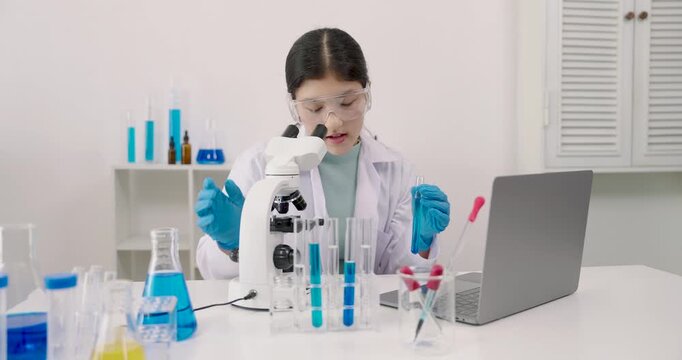 Asian girl participating in online science class using laptop while wearing safety goggles and gloves, holding testtube in hand surrounded by beakers and microscope on laboratory desk at school