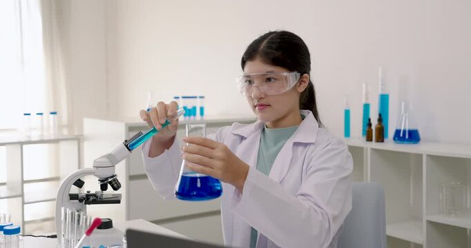 Asian primary student in labcoat using glass rod to stir chemical liquid in beaker during school science experiment, focusing carefully while sitting beside microscope and testtubes in laboratory