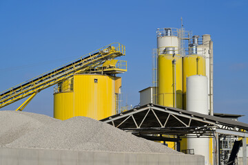 Obraz premium Industrial facility processing aggregates with conveyor belt and silos. Large industrial complex featuring silos, conveyor belts, and piles of raw material under a blue sky.