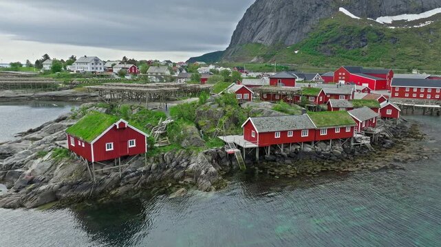 Aerial tracking shot over Hamn&oslash;y fishing village in Lofoten Islands, Norway. Norwegian red cabins and coastline framed by steep mountains and calm northern waters.