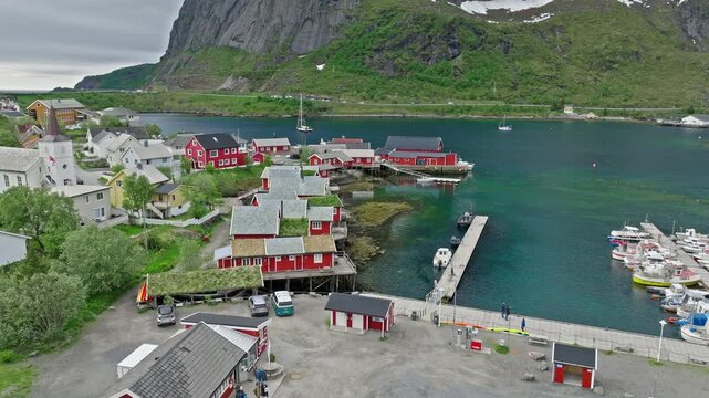 Drone aerial view of Hamn&oslash;y harbor in Lofoten Islands, Norway. Norwegian fishing village with traditional red cabins, pier and calm fjord waters beneath dramatic mountain landscape.
