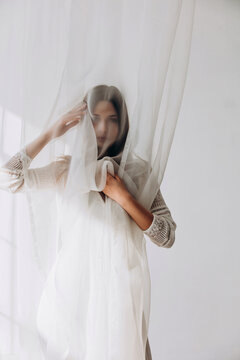 Ethereal brunette gently holding sheer white veil, soft dreamy portrait through translucent fabric.