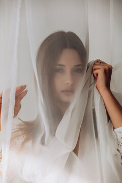 Ethereal brunette gently holding sheer white veil, soft dreamy portrait through translucent fabric.