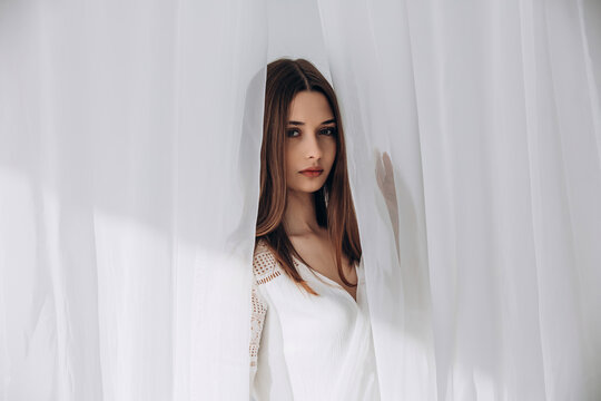 
Mysterious brunette peering through delicate white sheer curtains, soft ethereal portrait.