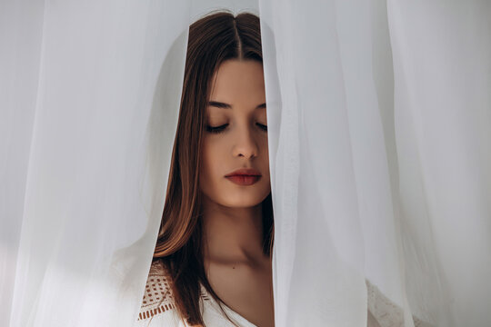 
Mysterious brunette peering through delicate white sheer curtains, soft ethereal portrait.