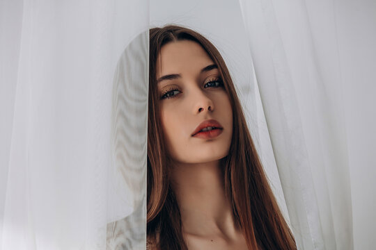 
Mysterious brunette peering through delicate white sheer curtains, soft ethereal portrait.