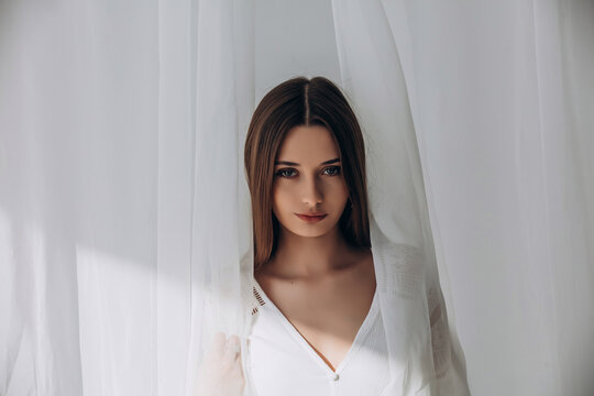 
Mysterious brunette peering through delicate white sheer curtains, soft ethereal portrait.