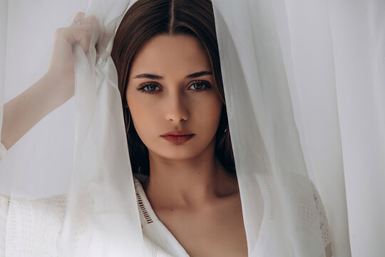 
Mysterious brunette peering through delicate white sheer curtains, soft ethereal portrait.
