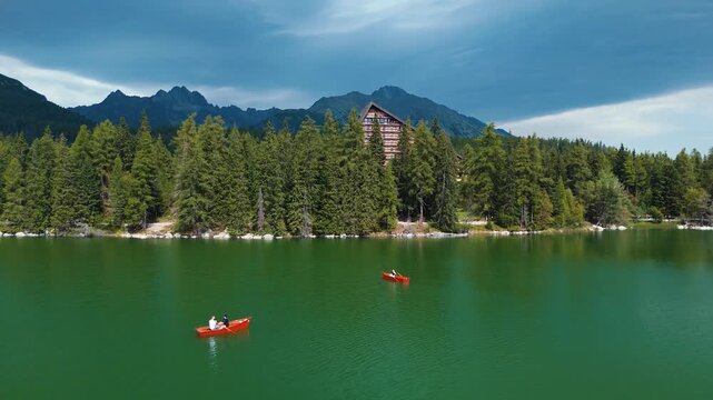 Drone view of rowboats on &Scaron;trbsk&eacute; Pleso in Slovakia with High Tatras mountains, forest shoreline and alpine hotel under dramatic sky.