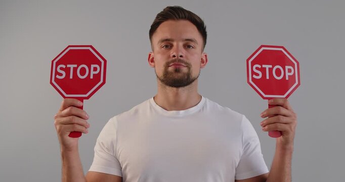 Confident man poses with a red octagonal stop sign, delivering a clear warning gesture against a plain grey backdrop. Clear stop signal for safety awareness. Prohibition and restrictions concept
