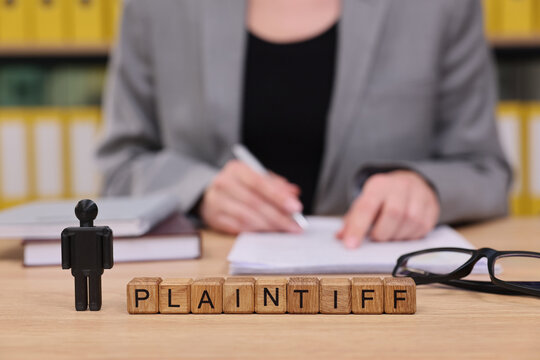 In a bustling legal office, a focused plaintiff stands beside wooden blocks spelling plaintiff, while a lawyer jots down notes. The atmosphere is charged with anticipation and seriousness.