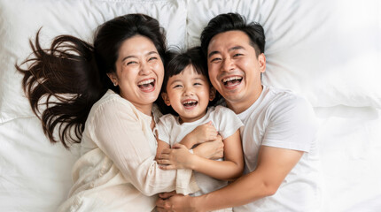 Obraz premium Joyful Asian family with young daughter laughing together on a white bed, overhead view