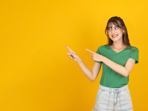 Cheerful young woman in green t-shirt pointing with both index fingers at copy space and smiling against vibrant yellow background. Concept of presentation, showing advertisement, and directing attent