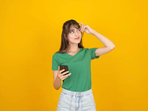 Thoughtful young woman in green t-shirt holding smartphone and scratching her head while looking away. Concept of forgetting a password, choosing an app, or searching for information against yellow ba
