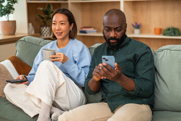 Obraz premium Multiracial couple relaxing at home. Woman is watching tv and man is using smartphone.