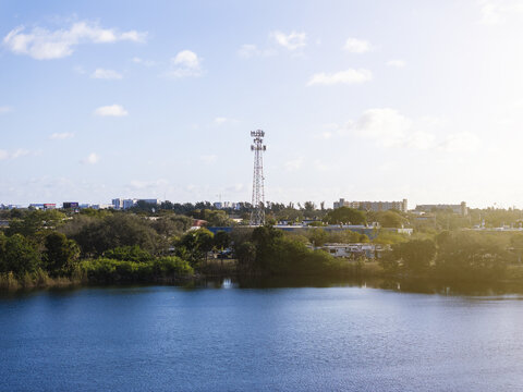 Aerial view of a cell phone tower rising above the cityscape, its stark metal contrasting with the lush greens and blues below, reflecting under a vast, open sky, Hollywood, Florida, United States.
