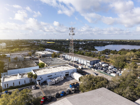 Aerial view of a cell phone tower rising above industrial buildings, nestled near a serene lake under a partly cloudy sky, Hollywood, Florida, United States.