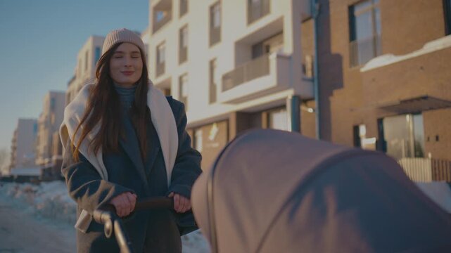 Caucasian woman pushing baby stroller. Wearing knitted hat and warm coat. Looking at child while walking slowly at winter street. Modern residential district. Caring mother enjoying walk outdoors.