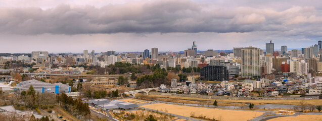 Obraz premium Panoramic view of Sendai from the historic Aoba Castle Ruins, overlooking a modern cityscape framed by lush greenery and distant mountains under a clear sky in Japan.