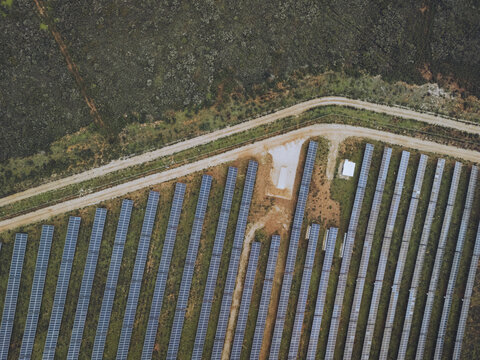 Aerial view of solar panels reflecting the sun's rays stand in orderly rows beside a sandy path, contrasting with the wild, untamed landscape, Lagos, Faro District, Portugal.
